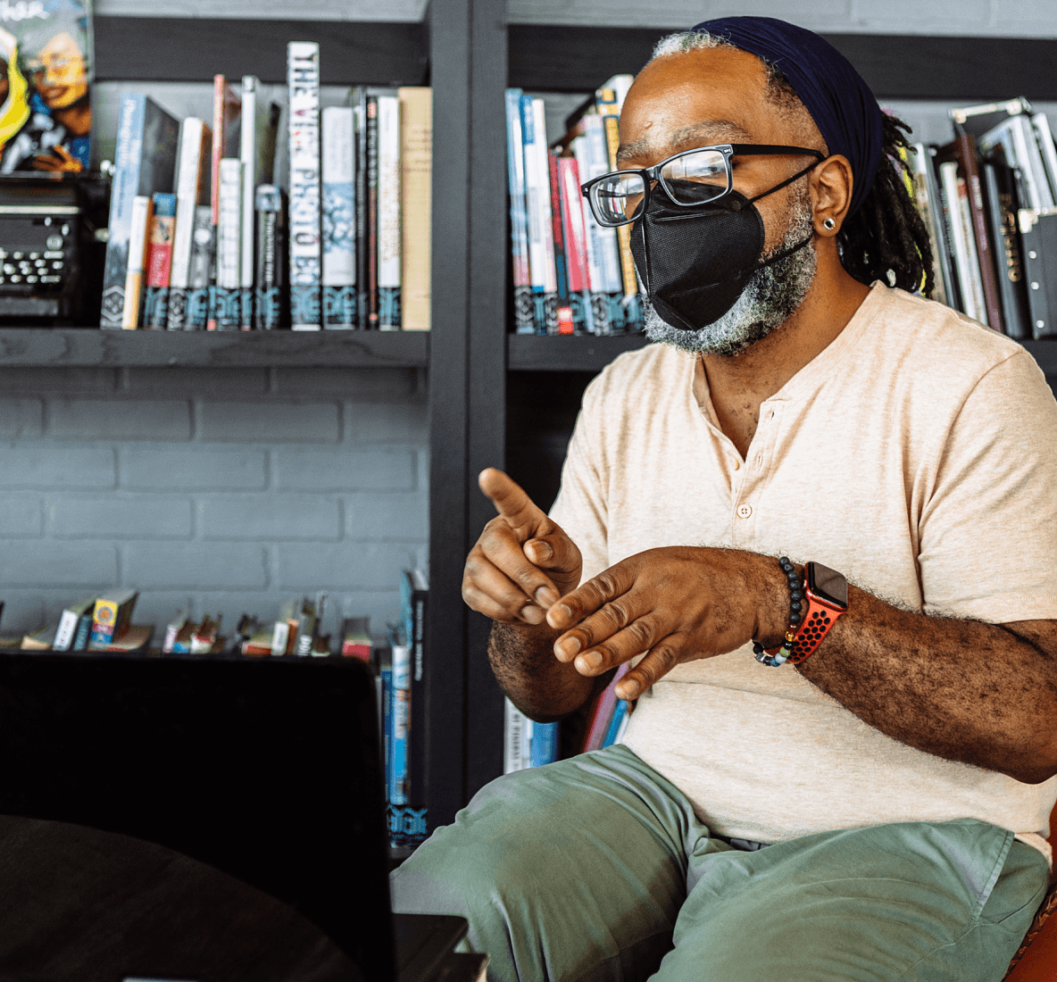 Ritratto di un uomo nero sordo che indossa occhiali e una mascherina KN-95 mentre comunica in una videochiamata tramite computer portatile. L'uomo è seduto a un tavolino da caffè all'interno di una biblioteca comunale piena di libri e poster.