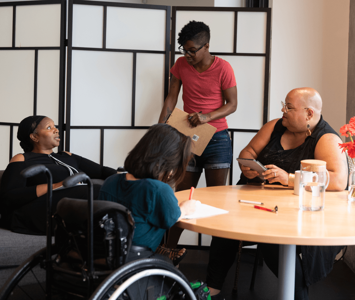 Quattro persone di colore con disabilità si riuniscono attorno a un tavolo durante una riunione. Una donna nera seduta su un divano parla con un'espressione neutra mentre le altre tre persone (una persona sudasiatica seduta su una sedia a rotelle che prende appunti, una persona nera non binaria seduta su una sedia con un tablet e un bastone e una persona nera non binaria in piedi con un blocco per appunti) ascoltano.